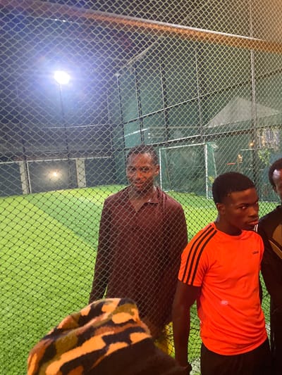 The image captures a group of young men on a fenced Astroturf field, likely at a sports facility in Malete, Nigeria, given the presence of the Kwara State University signage visible in the background. The scene is illuminated by artificial lights, casting a bluish-white glow over the bright green turf. In the foreground, the mesh of a chain-link fence partially obscures the view.

On the left, a man in a maroon collared shirt stands behind the fence, his face partially visible. To his right, a young man in a vibrant orange Adidas t-shirt with black stripes on the sleeves, and black shorts, is the central focus. He is looking intently towards the right of the frame. Another individual, wearing a black garment with gold accents, stands to the far right, his face obscured. In the extreme foreground, a person is seated, wearing a camouflage patterned garment, their face out of frame. The background reveals further fencing and hints of structures, typical of a sports complex. The overall atmosphere appears to be casual, possibly before or after a game or training session.