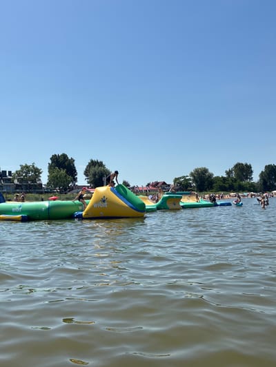 The image shows a bright, sunny day at a water park in Rewa, Poland. In the foreground, the calm, murky water of the Baltic Sea is visible, with gentle ripples reflecting the sunlight. In the middle ground, a series of large, inflatable Wibit water obstacles are set up. These are brightly colored, with green and yellow predominating. People are actively playing on these structures, with some climbing, jumping, and balancing. One person is visible on a yellow, pyramid-shaped obstacle, while another is on a long, green, cylindrical obstacle. In the background, the sandy shore of Rewa is lined with trees and buildings, indicating the proximity of the town. Beachgoers can be seen relaxing on the sand and swimming in the water. The sky is a clear, vibrant blue.
