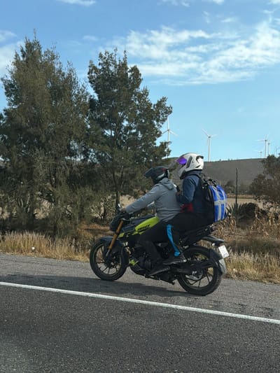 Two people on a motorcycle, one with a white helmet and the other with a black one. The bike is on a road with a white line. Behind them is a tree and a blue sky with some clouds. In the distance, there are a few wind turbines. The photo is taken outdoors.