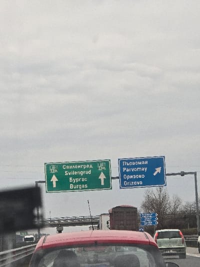 The image depicts a multi-lane highway scene in Bulgaria, specifically near the Orizovo exit. The perspective is from within a red passenger vehicle, whose rear roofline and antenna are visible in the lower foreground. Overhead, a gantry displays two main road signs: a green sign on the left indicates "Svilengrad" and "Burgas" with straight-ahead arrows for routes A1 and E80; a blue sign on the right directs traffic towards "Parvomay" and "Orizovo" via a right-pointing diagonal arrow, indicating an upcoming exit. Ahead of the red vehicle, traffic includes a light-colored passenger car with a partial Bulgarian license plate (e.g., "PZ 2707 PZ") in the right lane, and a large brown truck further ahead. A concrete overpass structure spans the highway in the background. Bare-branched trees line the highway's right side. The sky is overcast and uniformly grey. No individuals are visible outside of vehicles.