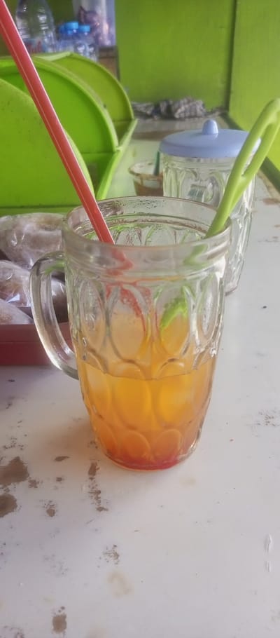 The image shows a glass of orange juice with a red straw and a green straw in it. The glass is sitting on a white table. There is a green plastic container in the background with a few items inside. The glass is sitting next to a second glass jar with a blue lid and a green straw. The image was taken in Kecamatan Waru, Indonesia. The coordinates are -7.35 latitude and 112.764 longitude.