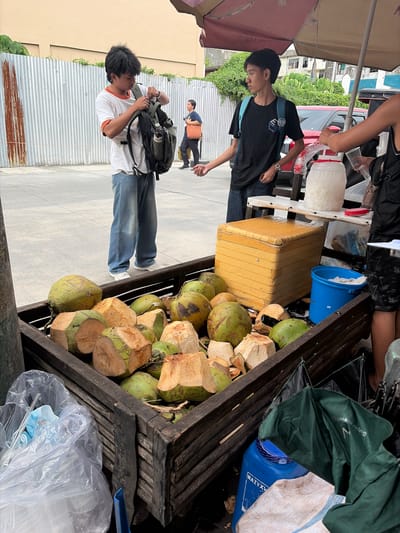 At a street-side vendor stall in Dagupan City, Philippines, a young male with dark, messy hair is adjusting the straps of a green backpack while wearing a white t-shirt with an orange trim and denim jeans. Behind him, a corrugated metal fence stands as a backdrop. To his right, another young male, with short dark hair, wearing a black t-shirt and a light blue backpack, is looking to his left. He stands near a vendor's cart filled with coconuts, some whole and others halved. A large yellow cooler sits on the cart, next to a clear plastic container holding a white substance, likely water or ice. A blue bucket and a plastic bag containing a blue mask and straws are also visible on the cart. The vendor is serving a customer, handing them a plastic cup. The ground is paved concrete, and a red vehicle is parked behind the vendor's stall. A large blue plastic drum is located at the base of the cart, next to a green tarp. The scene appears to be an outdoor market or roadside refreshment stand, with a red car and buildings visible in the background.