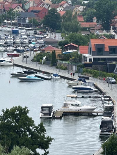 The image depicts a harbor in Mikołajki, Poland, on a cloudy day. The water is calm, with several boats docked along a paved walkway. The boats vary in size and type, including yachts, speedboats, and smaller motorboats. Some boats have canvas covers, while others are open. A few people are visible walking along the walkway, and one person is standing near a silver car.
In the background, a town with red-tiled roofs and multi-story buildings rises above the harbor. Lush green trees are interspersed among the buildings and along the shoreline. The overall scene is one of a well-maintained recreational harbor with a picturesque town backdrop.