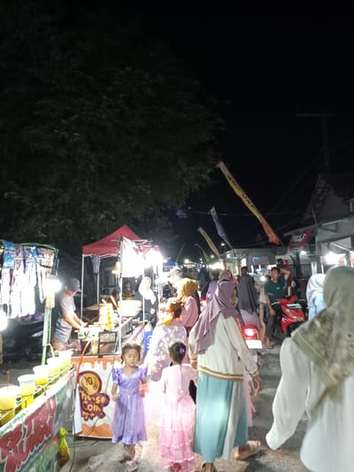 At night in Blitaran, Indonesia, a crowded street market buzzes with activity. Several people are seen in the foreground, including two young girls in dresses and women in hijabs walking through the market. Further back, vendors are working in food stalls under bright lights, while more people mill about. Banners stretch above the crowd, and a red scooter is visible amidst the people. Trees are partially visible in the darkness at the edge of the scene, with power lines and building roofs beyond.