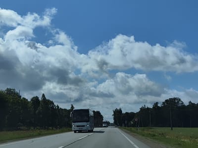 The image depicts a highway scene in Svētciems, Latvia. A white bus, identified by its headlights and license plate, is traveling on a paved road. The road is flanked by dense green trees and vegetation on both sides. In the distance, other vehicles, including a truck, are visible on the highway. The sky is blue with scattered white and gray clouds. Signs are present along the roadside, one of which indicates a speed limit of 70. The lighting suggests it is daytime.