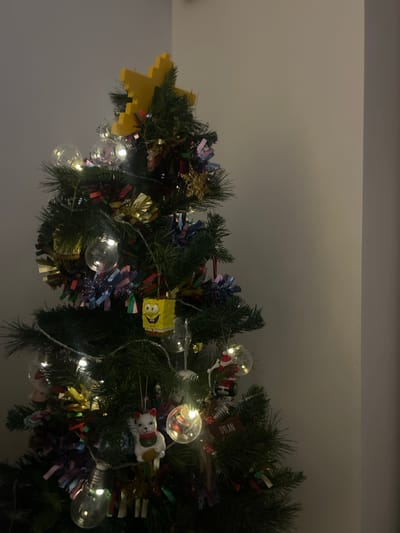 The image displays an artificial Christmas tree located indoors in Corrimal, Australia. The tree is topped with a pixelated yellow star and decorated with multi-colored tinsel. A string of warm white LED lights with clear, spherical bulbs illuminates the branches. Visible ornaments include a yellow Spongebob Squarepants figure, a white cat figure holding a gold object, a miniature Santa Claus figure, and a brown rectangular ornament inscribed with "DYLAN." The tree stands against a plain, light grey wall, which exhibits a corner on the right side of the frame. The scene is dimly lit, with the primary light source originating from the tree's decorations.