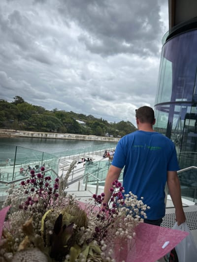 A bouquet of flowers with pink wrapping paper sits in the foreground. Behind it, a man in a blue shirt looks out at a gray, cloudy sky. The scene appears to be taken from a high vantage point, possibly a walkway or observation deck with glass barriers, as other people can be seen walking further along. Trees are visible in the mid-ground, creating a backdrop of natural greenery.