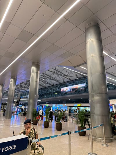 The image depicts the interior of an airport terminal, likely at Kempegowda International Airport Bengaluru, given the mention of Gollapalle Khurd, India, which is in the vicinity. A security guard, wearing a camouflage uniform and a beret, sits in a wheelchair in the foreground, looking towards the camera with his hand on his chin. Behind him, a stanchion with blue and white tape separates different areas of the terminal. In the mid-ground, several potted plants are strategically placed, and a line of people can be seen, some of whom appear to be queuing. Further in the background, various shops and food establishments are visible, including a Subway restaurant. The architecture of the terminal features large, cylindrical metallic pillars supporting a modern, multi-level ceiling with integrated lighting. The floor is polished and reflects the overhead lights and surroundings. The overall atmosphere suggests a busy transit hub.