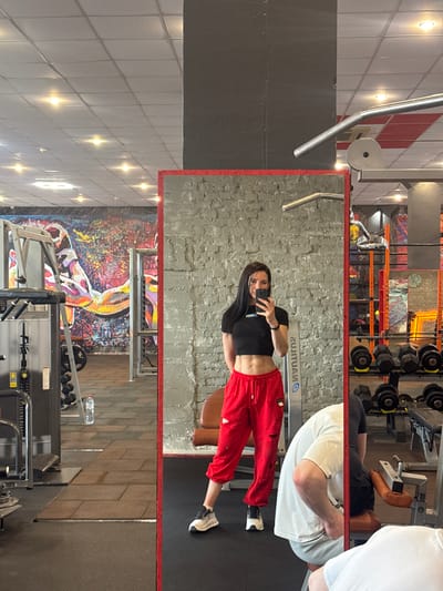 The image captures a reflection in a mirror, likely taken in a gym located in Assinovskaya, Russia. The primary subject is a young woman wearing a black crop top and red track pants, holding a smartphone and taking a selfie in the mirror. Behind her in the reflection is a textured gray wall.

The gym environment is visible both in the mirror and outside of it. Gym equipment, including weight racks, Nautilus machines, and other exercise apparatus, is discernible. Other individuals are also present in the gym, including a person in a white t-shirt and gray shorts, as well as another person using a seated machine in the background.

The gym’s interior features ceiling tiles with recessed lighting. A colorful painting adorns the wall below the mirror.
