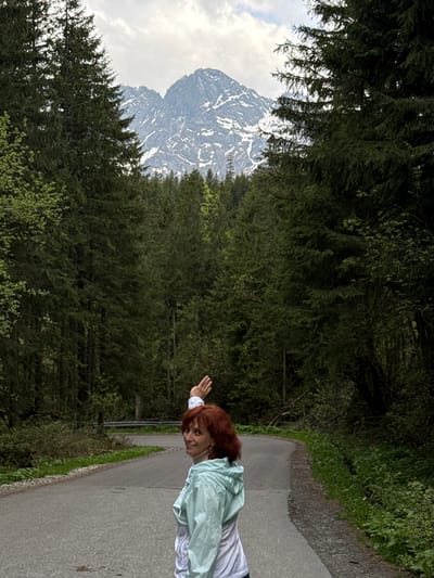 A woman stands on a paved road in a wooded area with mountains in the background. The woman has red hair and is wearing a white shirt and a light green jacket. She is extending her arm towards the left side of the frame. The road is bordered by a green embankment on the right and dense trees on the left. In the distance, a tall, snow-capped mountain is visible through the trees, set against a cloudy sky. The overall scene appears to be outdoors in a natural setting. The picture is rotated 90 degrees counter-clockwise. The location of Brzegi, Poland is not mentioned in the picture, and it is not apparent if the picture was taken there.