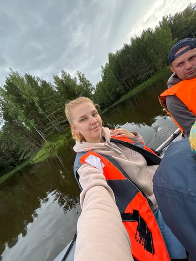 A selfie is taken from a boat on a lake, with a woman in the foreground and a man in the background. The woman, with blonde hair pulled back, is wearing a beige hoodie and an orange life vest. She is looking directly at the camera and smiling. The man, wearing a dark baseball cap and an orange life vest, is looking slightly off-camera to his right. The boat is moving on a dark, calm lake, with a dense forest lining the shore. The sky is overcast with gray clouds. The location is identified as Anteplevo (Okhotinskoe S/P), Russia.