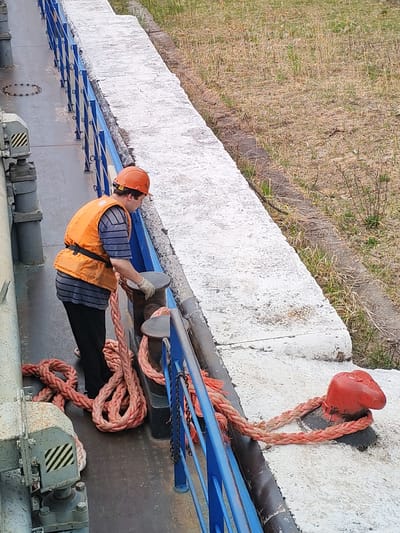 A worker in Alekseevskoe, Russia, secures a large rope to a bollard on a vessel. The worker wears an orange safety vest, a striped shirt, black pants, work gloves, and an orange hard hat. A blue railing borders the edge of the vessel. The shoreline is visible beyond a concrete barrier. The rope is thick, twisted, and reddish-orange. A second bollard is nearby on the vessel.