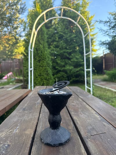 An outdoor scene features a weathered wooden picnic table in the foreground. A dark, ornamental holder with a lit mosquito coil sits centered on the table. In the background, a white garden archway stands amidst lush green trees and foliage. The sky above is a clear blue. The location appears to be a residential garden or park in Тмз Снт, Russia. There are no people visible in the frame.