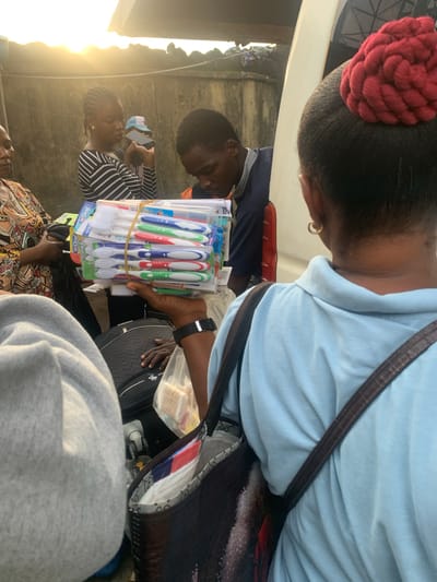 In Elelenwo, Nigeria, an outdoor scene depicts a group of people near a vehicle. A young woman with braided hair, wearing a black and white striped shirt, holds a smartphone and a blue and white cap. Beside her, a young man in a blue and orange shirt looks down. In the foreground, a person in a light blue shirt with a red braided hairstyle tied up on their head holds a stack of toothbrushes packaged in plastic. They are wearing a gold hoop earring and a dark brown crossbody bag. Another individual, seen from the back, wears a grey hooded garment. A large dark bag with the brand name "SWISS" visible is on the ground. The lighting suggests it is late afternoon or early morning, with the sun low in the sky. The background consists of a textured wall and a portion of a white vehicle.