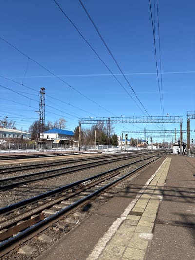 The image depicts a railway station in Kropachyovo, Russia, under a clear, bright blue sky with a single faint contrail visible. The scene is devoid of individuals. In the foreground, a concrete railway platform is visible on the right, featuring light-colored tactile paving blocks along its edge. Multiple sets of parallel railway tracks, approximately five visible, extend into the distance, with dark gravel ballast between them. Shadows from unseen structures and overhead lines are cast across the tracks and platform.
To the left of the tracks, in the midground and background, there are various buildings, including a structure with a distinctive bright blue roof and a multi-story building to its left. Bare trees and sparse brush are present behind a fence separating the tracks from these buildings. Patches of snow are visible on the ground beside the tracks and near the buildings, indicating a late winter or early spring setting. Overhead, a complex network of electrical catenary lines is suspended by metal gantries and poles. A tall, brown, lattice-style metal tower stands to the left of the tracks, possibly supporting signals or communication equipment. Further along the tracks on the right, a small white building, likely a signal box or utility structure, is present near the end of the platform. Several railway signals with dark casings and circular lenses are visible along the track lines in the distance.