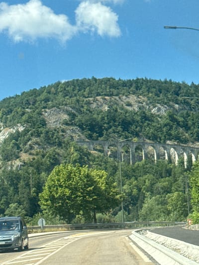 The image captures a scenic view of a mountainous landscape, likely in the region of Hauts de Bienne, France. In the foreground, a gray vehicle, possibly a van, is seen driving on a paved road. The road curves to the right, bordered by a concrete barrier on the right side. In the middle ground, a distinctive stone aqueduct with multiple arches traverses the hillside. The aqueduct is surrounded by lush green trees. The background is dominated by a steep, tree-covered mountain, with rocky outcrops visible. The sky above is blue with scattered white clouds, and a street lamp is visible on the right side of the frame. The overall impression is one of a natural, somewhat rugged environment integrated with historical infrastructure.