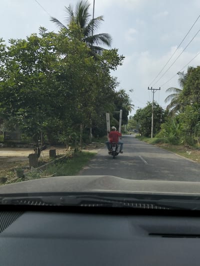 The image, captured on a road in Blang Mee, Indonesia, shows a person riding a motorcycle down a paved road lined with lush green trees and vegetation. The rider, wearing a red shirt and blue jeans, is carrying two long, vertical white poles on the back of the motorcycle. The sky is partly cloudy. In the foreground, the dashboard of a vehicle is visible, suggesting the photo was taken from inside a car. The scene depicts a common mode of transportation and cargo carrying in the region.