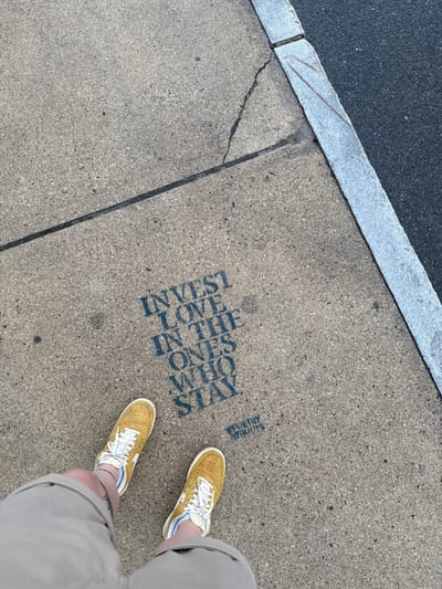 The scene captures a sidewalk in Hampton, United States, featuring a person's feet in tan sneakers and tan shorts. Etched onto the concrete of the sidewalk are vertically stacked words: "INVEST LOVE IN THE ONES WHO STAY." A strip of asphalt is present at the top left, bordered by a cement curb. A thin crack runs along the sidewalk surface near the curb. The writing includes "RICH IN ROOTS" just above the letter "S".