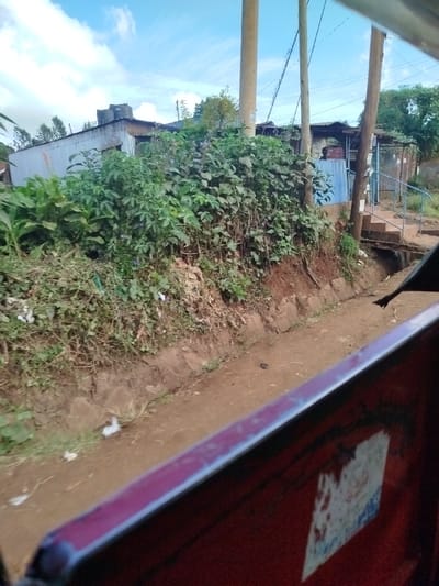 The image displays an outdoor scene in Kihara ward, Kenya, captured from a vehicle, with a red-colored interior frame partially visible in the bottom right foreground. An unpaved, light brown dirt path traverses the midground. To the left of this path, a steep earthen embankment is covered with dense green vegetation and scattered refuse. A narrow, concrete-lined drainage channel runs parallel to the path, with a small bridge or wooden plank crossing it. In the background, several structures constructed from corrugated metal are visible, including one with two black water tanks on its roof. Three utility poles with power lines extend vertically through the scene. A blue corrugated metal structure is visible near the center poles, and a set of concrete steps with blue metal railings ascends from the path on the right side. The sky above is partly cloudy with blue patches. No individuals are present in the frame.