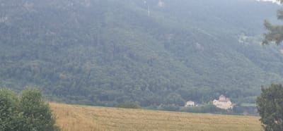 The image displays a landscape with a castle and a hill in Rans, Switzerland. The castle, a prominent structure, is situated on a hill. The foreground features a field of dry grass, indicating a rural or natural environment. The background is dominated by a densely forested mountain. The overall lighting suggests an overcast or hazy day, with soft light casting over the scene. The image is a static, wide shot, capturing the natural beauty and architectural elements of the location. There are no people or discernible actions taking place in the image.