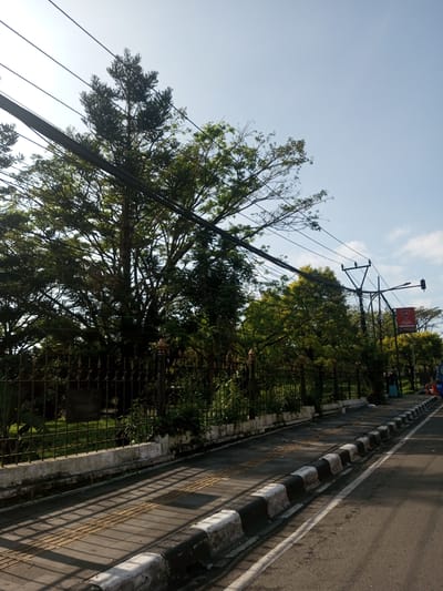 The image depicts a street scene in Nhot Chang, Laos. The view is from the street level, looking towards a green space bordered by a low concrete wall topped with a dark metal fence. A row of mature trees lines the fence, partially obscuring the park behind it. Utility poles and wires run along the side of the street. The street itself has a curb painted in black and white stripes, separating the road from the sidewalk. A portion of a traffic sign is visible further down the street. The sky is clear with some scattered clouds. A pedestrian in a long dress and a parked vehicle are visible in the distance.
