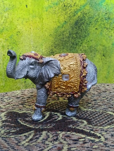 A miniature, silver-colored elephant figurine with ornate gold and red decorative coverings stands on a patterned surface against a blurred, variegated green background. The elephant is depicted with its trunk raised. The figurine exhibits intricate detailing on its skin and embellishments. The patterned surface appears to be a textile. The background is abstract and out of focus, suggesting it is not a primary element of the scene. There are no people or active interactions depicted. The image captures a static, posed subject. The location in Florești, Moldova is not discernible from the visual information present in the image.