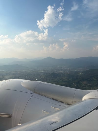 An aerial view taken from an airplane window shows a hazy landscape of rolling hills and a densely populated city or town below. Mountains are visible in the background, receding into the distance under a partly cloudy sky. The foreground is dominated by the wing and engine nacelle of the aircraft. The wing is metallic and appears to be the leading edge, with rivets visible. The engine nacelle is large and cylindrical, with a smooth, reflective surface. The lighting suggests it is daytime, possibly late afternoon, given the soft, diffused light and the haze. The image appears to have been taken in the vicinity of Orio al Serio, Italy, based on the mountainous terrain that is characteristic of the region surrounding Bergamo, where Orio al Serio is located. No people or specific actions are discernible in the image.