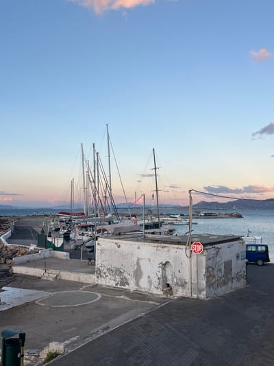The image captures a harbor scene at either dusk or dawn within the Megalochori, Greece area. In the foreground, a dilapidated, single-story rectangular building with weathered, peeling white stucco and an exposed, flat roof occupies the right. A red circular "STOP" sign is mounted on a pole directly in front of this structure. To the immediate right of the building, a blue utility van or similar vehicle is parked. The ground consists of concrete paving stones and rougher concrete slabs. In the bottom left, a green waste bin is partially visible. The midground features a marina populated by numerous moored boats, predominantly sailboats characterized by multiple tall masts, along with several motorboats. One sailboat prominently displays the partial text "NEXUS" on its hull. The water is calm and reflects the sky. In the background, a distant landmass with rolling hills is visible. The sky transitions from a light blue overhead to soft pink and orange hues near the horizon, interspersed with wispy clouds. A small lighthouse or navigational beacon is situated on a distant pier or breakwater to the far right. No people are present in the scene.