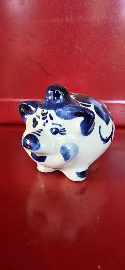 A ceramic pig figurine, painted with blue floral patterns on a white background, sits on a shiny red surface. The figurine is facing left, with its snout slightly elevated. The background is a solid red color with some subtle texture. The lighting casts a reflection of the figurine onto the red surface below. The scene appears to be indoors, given the lighting and the smooth, uniform background. The location is not specified in the image itself.