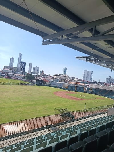 The image displays a panoramic view of a baseball stadium, identified as being in Curundú, Panama, observed from an elevated seating area. In the immediate foreground, rows of unoccupied, dark green plastic stadium seats are visible, positioned beneath a modern, shaded roof structure composed of dark structural beams and light-colored panels. A dark metal barrier separates the spectator area from the field.
The midground features a verdant green grass outfield and a reddish-brown dirt infield, characteristic of a baseball diamond. A green, mesh batting cage or protective net rests on the dirt near the first base foul line. Beyond the playing field, a section of older, concrete stadium grandstands with a more traditional roof is visible. Further in the background, a dense urban landscape unfolds, comprising numerous low-rise buildings and what appears to be a busy street with visible vehicles.
The distant background is dominated by a clear, bright blue sky and a distinctive skyline of modern skyscrapers, indicative of Panama City's urban core. No individuals are present in the image, and no active events or interactions are depicted. The scene is illuminated by strong sunlight.