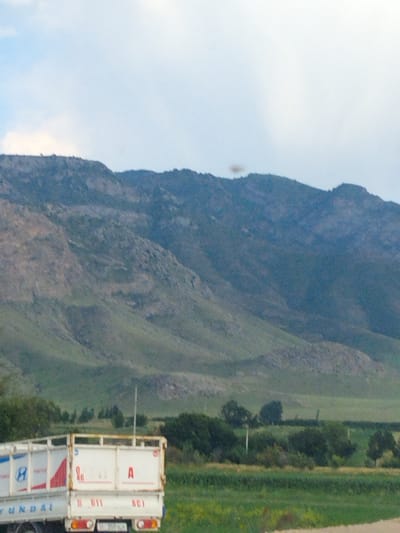 A white truck is parked in front of a mountain range. The truck has a blue logo with the letter H inside a circle on its side. There are also red and white stripes on the truck. The mountains are covered in green grass and there are some trees in the foreground. The sky is blue with some clouds. The location is the city of Jerge-Tal, Kyrgyzstan.