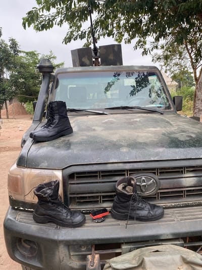 The image displays a dark green, dusty, modified Toyota Land Cruiser-type vehicle parked on dirt ground in Kado, Nigeria. The vehicle's front section is prominent, featuring a snorkel on its left side and a visible Toyota emblem on the front grille. A heavy machine gun, flanked by two rectangular dark armored plates, is mounted on the vehicle's roof, positioned behind the windshield.
A pair of new-appearing black laced combat boots rests on the vehicle's hood. On the front bumper/grille, a second pair of visibly worn black laced combat boots is present, alongside a circular black shoe polish container and a red and black shoe brush.
The background consists of sparse green trees and vegetation, a large pile of reddish-brown earth to the left, and a partially obscured light-colored building in the distance. The sky overhead is overcast or hazy. No individuals are visible within the frame. The arrangement of boots and cleaning supplies suggests recent or impending maintenance activity.