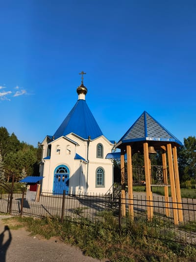 In the city of Aya, Russia, an Orthodox church with a white facade and a bright blue roof stands under a clear blue sky. A golden dome topped with a cross crowns the church's main structure. Adjacent to the church is a wooden bell tower with a similar blue roof and a bell hanging within. A black metal fence encloses the church grounds, separating it from a paved walkway and grassy area in the foreground. The scene is bathed in the warm light of what appears to be late afternoon or early morning, casting long shadows. A silhouette of a person holding a phone is visible in the lower left corner, suggesting the photo was taken by a visitor. Trees and foliage surround the church, indicating a semi-rural or suburban setting.