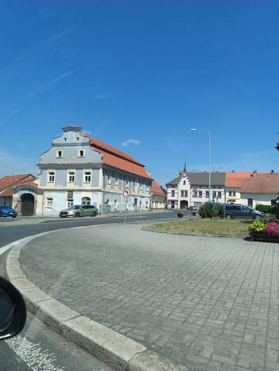 The image shows a street scene in Horažďovice, Czechia, on a sunny day with a clear blue sky. In the foreground, a curved road paved with grey interlocking bricks leads into a roundabout or intersection. A dark-colored car's side mirror is visible on the left, reflecting a portion of the scene.
On the right side of the road, a patch of grass with flower boxes filled with pink and green plants is visible. A grey van is parked on the right. Further down the street, a light pole stands tall. Behind the grass area, a building with a red tiled roof and white walls is visible.
To the left of the road, a large, pale blue building with a distinctive stepped gable and a red tiled roof dominates the scene. It has multiple windows and an arched entranceway with dark wooden doors. A blue sedan is parked to the left of this building. Adjacent to the blue building is another building with a red tiled roof and a light yellow facade.
Further down the street, a pink building with a stepped gable and a white tower-like structure on top is visible, along with other buildings featuring red tiled roofs. Traffic signs are present, including a "no left turn" sign with a red circle and a diagonal red line. The overall impression is of a quiet, European town square or street.