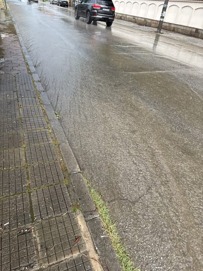 This image depicts a wet urban street scene in Parvenets, Plovdiv Province, Bulgaria, captured at an oblique angle looking forward along the roadway. The asphalt road surface is thoroughly saturated, exhibiting clear patterns of water flow and accumulation, particularly towards the left-hand curb.

A dark grey Jeep Grand Cherokee, bearing Bulgarian license plate PB 6178 HE, is positioned in the mid-ground on the right side of the road, with its rear section visible as it moves away from the viewer. Several other vehicles are discernible further down the street.

To the left, a concrete sidewalk, composed of square, textured tiles, is present. Green vegetation and moss are growing in the grout lines between the tiles and along the adjacent grey concrete curb. Minor organic debris, including leaves and small twigs, is scattered across the sidewalk surface.

On the right side of the street, a prominent white wall featuring a series of evenly spaced arched recesses runs parallel to the road. A dark utility pole, with affixed signage, stands near the right curb, casting a subtle reflection on the wet ground. The overall environmental conditions suggest recent or ongoing precipitation, with diffuse overhead lighting. No people are visible in the frame.
