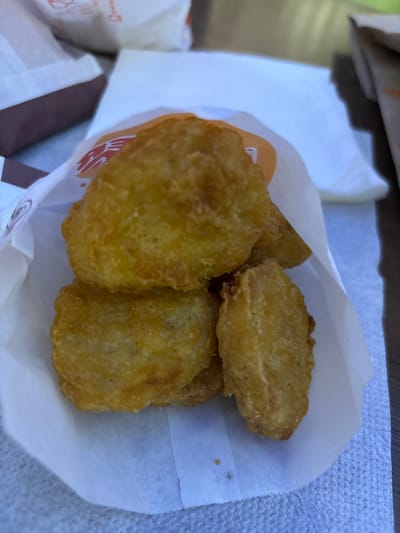 The image shows a close-up view of several golden-brown, crispy chicken nuggets nestled inside a white paper bag. The bag appears to be partially opened, revealing the food. The nuggets have a textured, breaded exterior. In the background, other paper food packaging can be seen, some with orange and red branding. The scene is lit by natural light, casting subtle shadows. The surface beneath the bag appears to be a textured, light-colored fabric, possibly a napkin or tablecloth. No specific location in the city of Shendzhii, Russia is identifiable from the image.