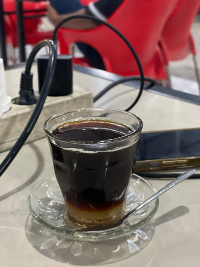 The scene is captured indoors in the city of Lhok Mereubo, Indonesia. A clear glass cup containing a layered beverage, specifically a dark liquid over a lighter, opaque cream-colored layer, is positioned centrally on a clear glass saucer with a small silver spoon. This assembly rests on a light grey table surface. To the left, a grey power strip has a black power adapter plugged into it, with a black cable extending from the adapter. To the right of the cup, a dark-colored smartphone with a light-colored bezel is lying flat on the table. In the out-of-focus background, parts of red plastic chairs and a person's leg dressed in dark blue fabric are discernible, suggesting a cafe or similar establishment.