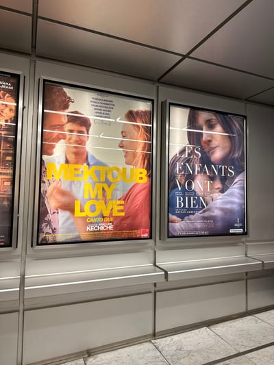 Two vertically stacked movie posters are visible. The top poster, in French, features a woman and baby, titled "LES ENFANTS VONT BIEN" starring Camille Cottin. The bottom poster, titled "MEKTOUB MY LOVE," in English and Italian, features actors Shain Boumedine, Jessica Pennington, and others. It's directed by Abdellatif Kechiche. Part of a third poster is barely visible at the bottom. The posters are displayed in black frames, against a wall with white paneling.