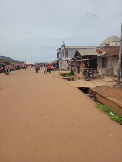 This is a photograph of a street scene in Bokkos, Nigeria, on an overcast day. The road is made of reddish-brown earth and appears to be dusty. Several motorcycles are present, some ridden by people and others parked. On the right side of the road, there are buildings with corrugated iron roofs and painted walls. One building has signage that reads "WHOLESALES & RETAILS". A satellite dish is visible on one of the roofs. A drainage channel runs along the side of the road, with some greenery growing in and around it. People are walking and standing in small groups near the buildings and on the road. The overall impression is of a busy, bustling local market or commercial area within the town.