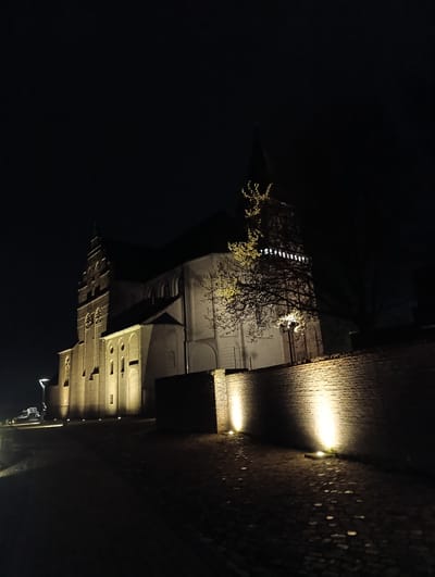 The image shows a night view of the Aldegundiskirche in Emmerich on the Rhine, Germany. The church and an adjacent brick wall are illuminated by ground-level lights. The church is a large, multi-story structure with pointed arches and a tower. A tree is partially visible in front of one section of the church. The ground is paved with stones and is dimly lit. No people are visible.