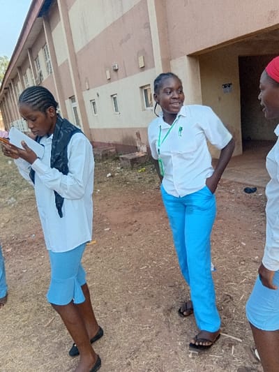 Three young women in school uniforms stand outside a light tan building. The uniforms consist of light blue bottoms and white collared shirts. One woman holds a phone. Another is standing and smiling, with her hand on her hip. Their hair is braided. The ground is dry, with some vegetation visible in the background. They are wearing sandals.
