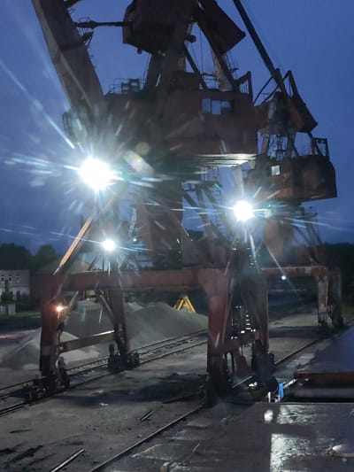The image depicts a large, weathered industrial gantry crane at night or late dusk/early dawn in what appears to be an industrial port or railway yard in Medvezhyegorsk, Russia. The dominant subject is the crane, a substantial structure made of rusty, dark-colored steel beams, featuring multiple support legs, cross-braces, and operator cabins.

Bright artificial lights are mounted on various parts of the crane, casting intense illumination and producing prominent star-shaped lens flares that pierce the dark blue sky. The sky itself shows faint cloud patterns. Below the crane, several parallel railway tracks are visible on a dark, possibly wet, ground surface that reflects some of the artificial light.

In the background, behind the crane's supports, are large piles of light-colored aggregate, such as gravel or sand. A piece of yellow heavy machinery, possibly an excavator or loader, is partially visible behind one of these piles. To the far left, a multi-story building with several windows can be seen against the horizon, partially obscured by trees or foliage.

No direct human activity is apparent, suggesting the crane is stationary or awaiting operation. The scene conveys a stark, industrial atmosphere. The rusty appearance of the crane indicates its age and consistent use in a demanding environment. No discernible text is visible on any objects within the photograph.