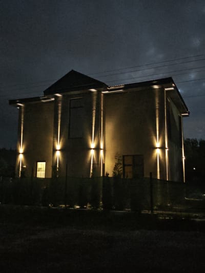 The image depicts a modern, two-story building with a dark, overcast sky. The building is illuminated by vertical, upward-facing wall lights that cast strong beams of light onto its beige exterior. There are several windows visible, with one on the lower left side glowing from within. A dark fence runs along the front of the property, with some dark green shrubbery behind it. The ground in front of the fence is dark and appears to be grass. In the background, dark trees are visible. Overhead, power lines are strung across the sky. The overall mood is somewhat dramatic due to the strong lighting and dark, cloudy sky. It appears to be nighttime. The location is identified as Надежда Снт, Russia. There is no text visible in the image.