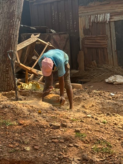 The image depicts an outdoor scene in Aguata, Nigeria. A single adult male, wearing a light blue short-sleeved shirt, dark shorts, and a pink baseball cap, is positioned centrally, bent over while holding a light-colored bucket filled with sand. He is barefoot, and a red band is visible on his right wrist. To the left, a dark-colored shovel leans against the trunk of a large tree. The foreground and midground consist of sandy, rocky soil with minimal sparse green vegetation. In the background, a dismantled wooden structure is visible near a dilapidated building constructed from corrugated metal sheets. Text painted on the metal building includes "STOP URINATING HERE" repeated multiple times. Other objects in the background include dried palm fronds, a white bag, and a tire. The scene is illuminated by daylight.