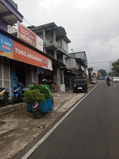 The image displays a daytime street scene under an overcast sky. In the foreground on the left, a concrete sidewalk is visible alongside an asphalt road. A white scooter is positioned with its front wheel on the sidewalk and rear on the road, heavily laden with a large volume of green leafy produce secured to its rear and two prominent blue fabric bags on its sides. A person wearing a dark helmet and a green long-sleeved garment is seated on this scooter, largely obscured by the produce. Behind it, a blue scooter is parked fully on the sidewalk.

Immediately above these vehicles, the ground floor of a commercial building features an orange banner with the text "TOKO AROHMAH" in white letters, and a smaller blue banner advertising "1 Liter Praktis." Above this, a large rectangular billboard displays the phrase "SEDAPNYA FAVORIT SEMUA" along with images of instant noodle products. The building is multi-storied, predominantly grey with white window frames and multiple balconies. Further down the sidewalk, a black pickup truck is parked, facing away from the viewer, with a white sack resting on the ground nearby.

The asphalt road, marked by a solid white line on its immediate edge, extends into the midground and background, showing multiple vehicles. Cars, including white and silver sedans, and additional motorcycles are observed traveling or parked. Buildings of varied architecture and height line the street into the distance, with some trees visible further back. Overhead power lines traverse the upper portion of the scene. A large, plain billboard stands in the far upper right background. The prominent signage on the buildings is in the Indonesian language.