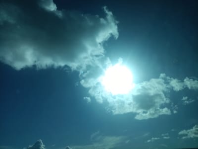 The image depicts a bright sun partially obscured by clouds in a clear blue sky, suggesting an outdoor scene in Třemošnice, Czechia. The sun is positioned to the right of the frame, casting a strong glare. Several large, white, and gray clouds are scattered across the sky, with some appearing more wispy and others denser. The bottom of the image shows a hint of a dark, indistinct horizon line, possibly indicating trees or distant structures. There are no visible people or animals in the frame, and no specific objects or actions are discernible beyond the natural elements of the sky. The overall impression is one of a bright, possibly sunny day with a mix of clouds.