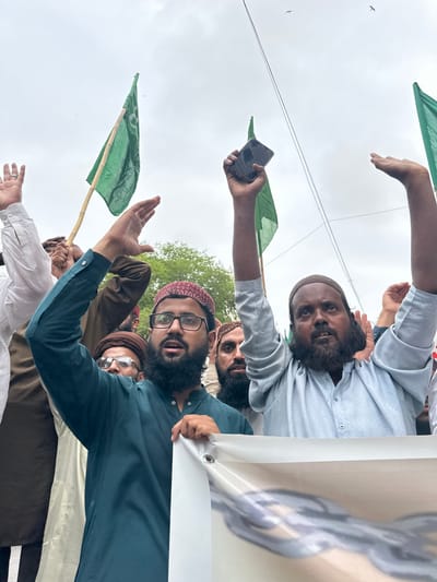 In Karachi Division, Pakistan, a group of men are participating in a demonstration. The man in the foreground wears a dark teal shalwar kameez and a maroon kufi, with glasses and a beard. He is raising his right hand. To his left, another man in a white shirt is also raising his hand. Behind them, a man in a light blue shalwar kameez with a beard is holding a mobile phone in his raised left hand and looking upwards. Several green flags are visible in the background. A white banner with a gray design is being held at the bottom of the image. The scene appears to be outdoors, under an overcast sky.