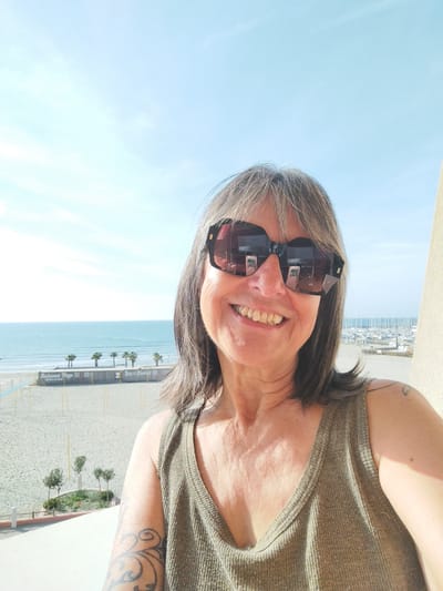 A medium-shot photograph captures a middle-aged woman smiling into the camera, wearing large sunglasses. She has graying hair and a light olive tank top. Tattoos are visible on her left shoulder and arm.

The background reveals a bright, sunny day in Palavas-les-Flots, France. A beach stretches out behind her, with the ocean visible in the distance. Palm trees line the beach. A structure labeled "Restaurant Plage" and "Bar & Cocktail" is also visible. A marina with many boats is seen on the right side of the frame, closer to the horizon. The photograph appears to be taken from a balcony.