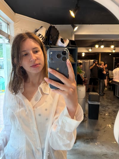 A young woman with brown hair is taking a selfie in what appears to be a retail or salon environment. She is wearing a white, sheer, button-down shirt over a darker garment. She is holding a smartphone in her right hand, which is capturing the image. The background is blurred but shows shelves with bags, a window to the left, and several people standing and interacting in the distance. The floor appears to be wet or reflective. The location is not explicitly identifiable as Seo-myeon, South Korea from the visual information provided in the image.