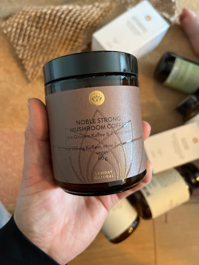 A light-skinned left human hand is holding a dark amber glass jar with a black screw-top lid in an indoor setting in Waldgrehweiler, Germany. The jar's brown label is centered, displaying the text "NOBLE STRONG MUSHROOM COFFEE," "mit Grünem Kaffee & 7 Vitalpilzen," "mit 120 mg Koffein, ohne Zucker," "vegan," "80 g," and "SUNDAY NATURAL." A gold lotus-like emblem is visible at the top of the label. The hand's thumb is positioned on the left side of the jar. In the blurred background, a light brown corrugated packing material is visible to the upper left. To the right, several other out-of-focus dark amber glass bottles and white cardboard boxes are present on a light-colored surface, consistent with a wooden tabletop.